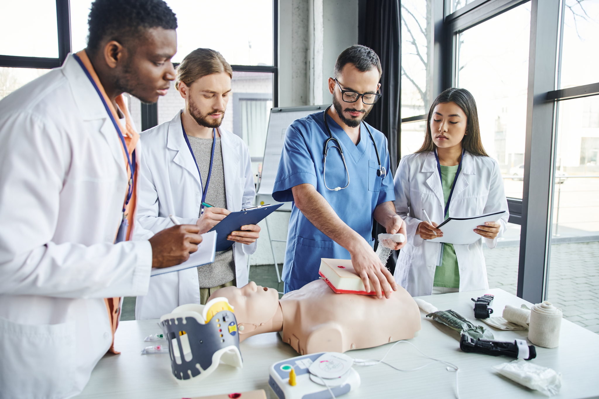 Medical students practice CPR on a manikin in a bright room. Diverse group focused, one in blue scrubs demonstrates chest compressions.