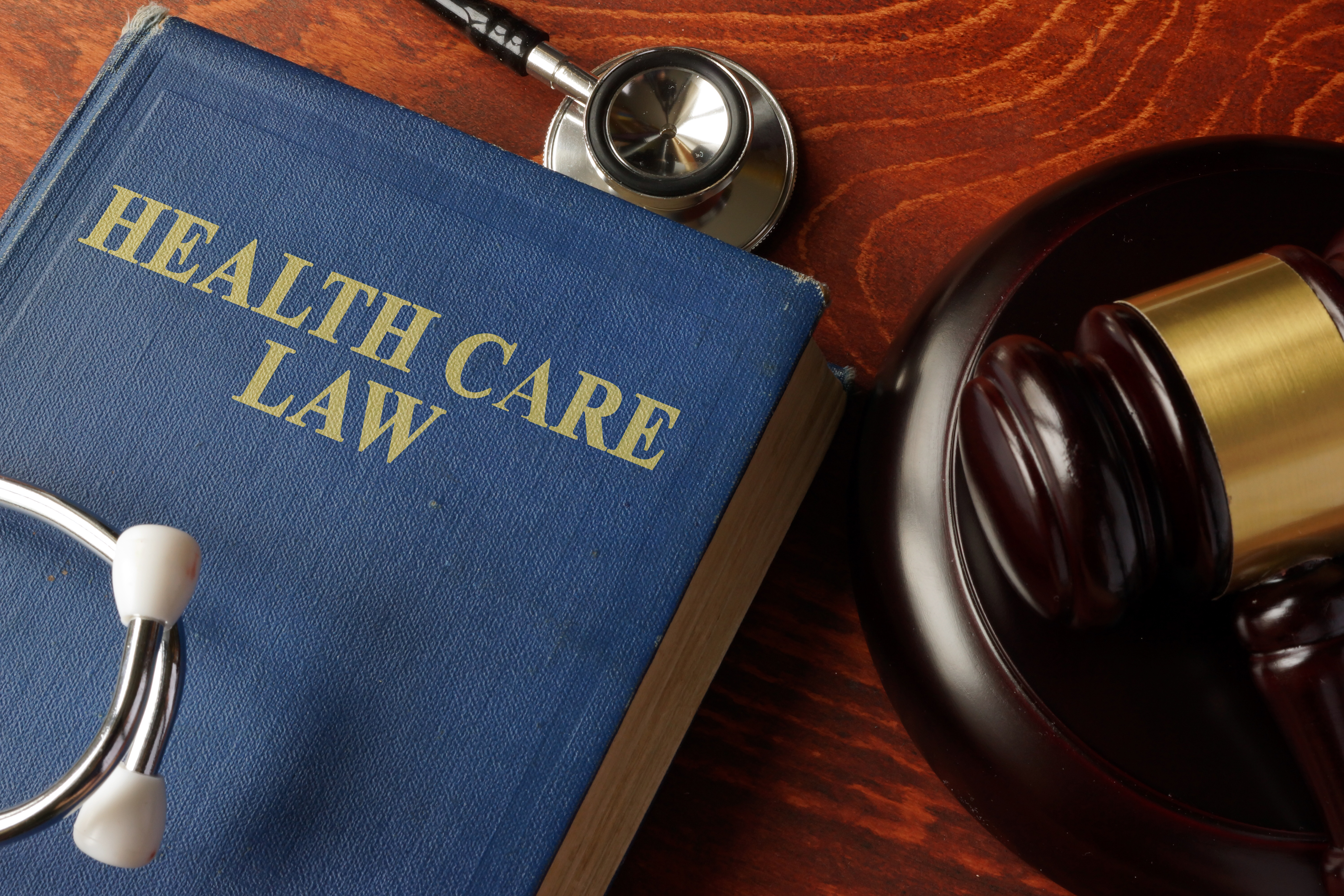 A blue book titled 'Health Care Law' lies on a wooden table beside a gavel and a stethoscope, symbolizing the intersection of health and law.